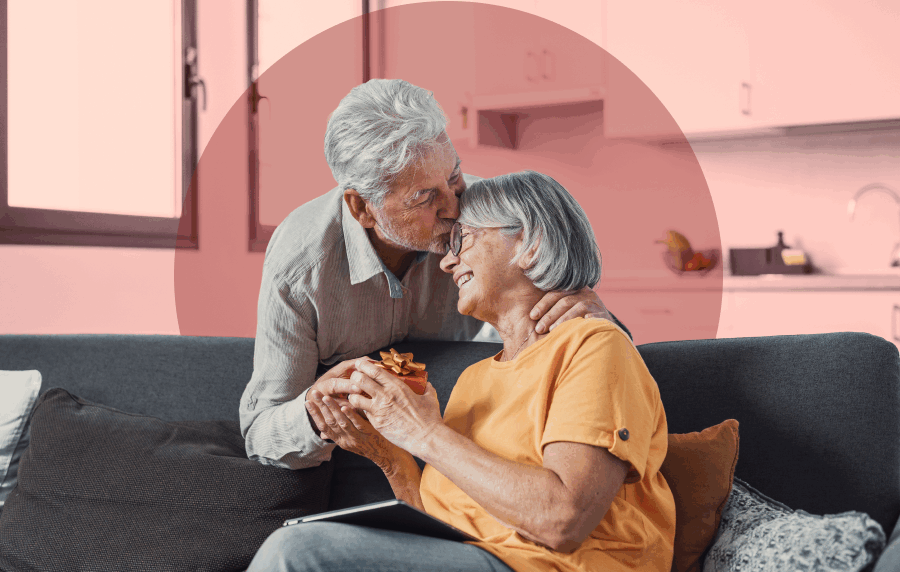 White senior couple in living room, the man bending over the couch where his wife is sitting, giving her a small wrapped gift and a kiss on the forehead