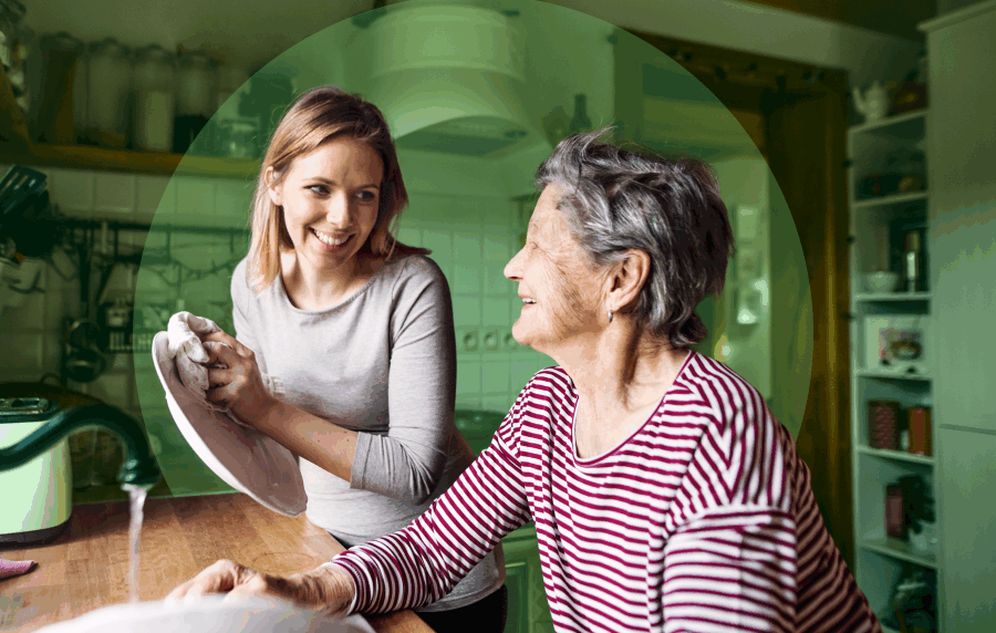 Elderly woman with grey hair and young blonde woman washing dishes together in a home kitchen, smiling