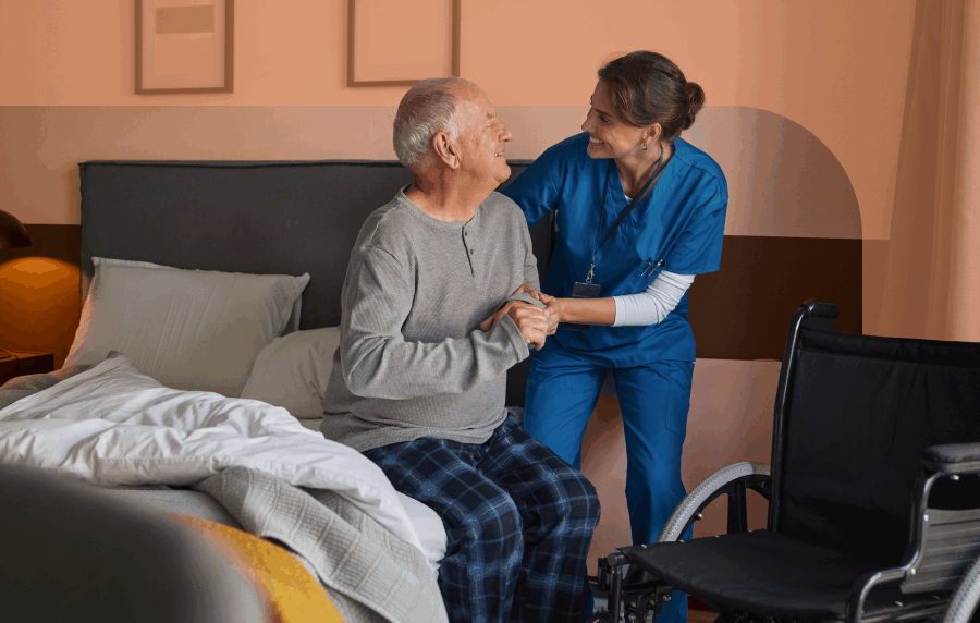 Senior man sitting at the edge of a bed smiling at a female nurse in scrubs who is assisting him. A wheelchair is in the corner.