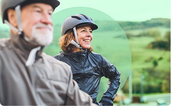 Two cyclists wearing helmets smiling outdoors with green landscape background