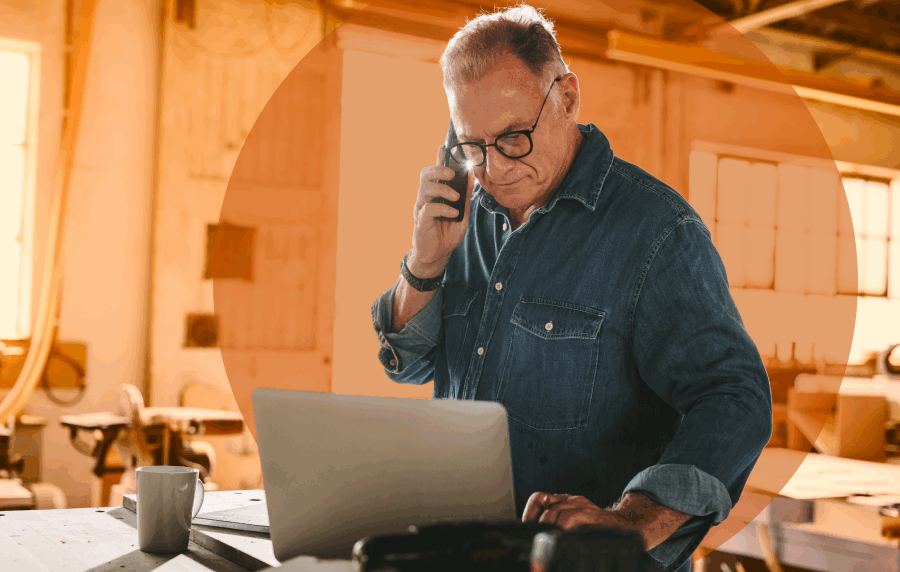 Senior man in a workshop wearing a denim shirt and glasses, holding a phone to his ear while looking at a laptop screen