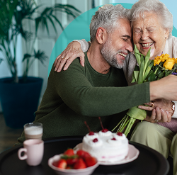 Elderly woman and man hugging, holding yellow flowers, smiling warmly.