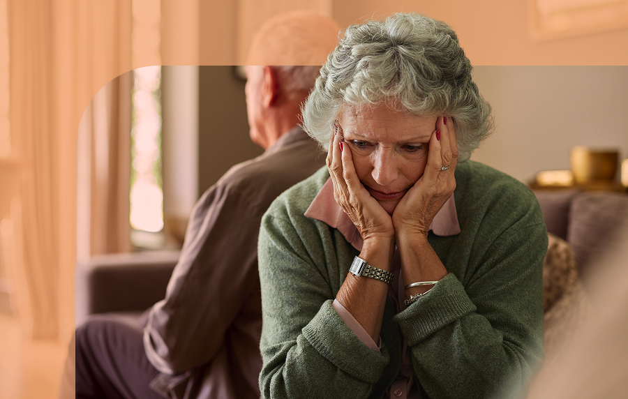 Senior woman in foreground, looking distressed with her hands cupping her face. A senior man is sitting facing away from her in the background.