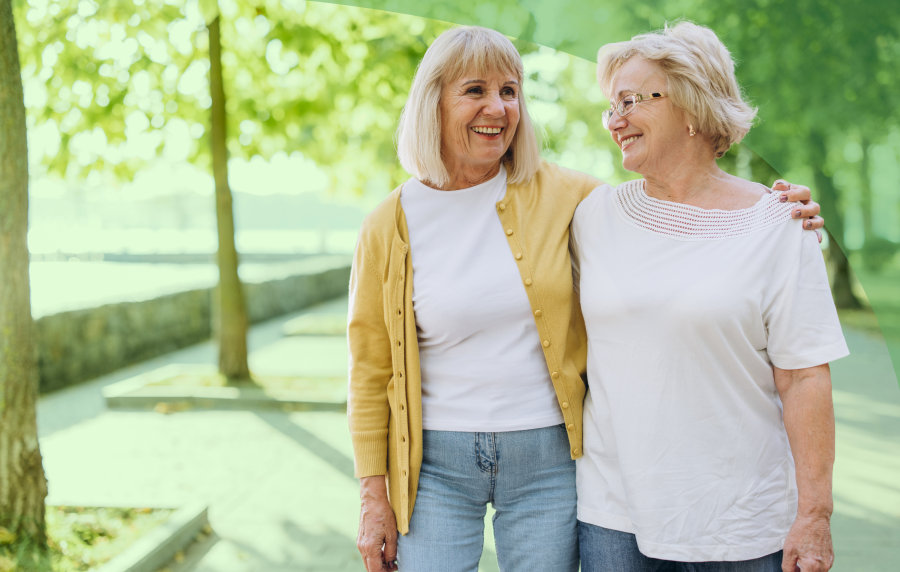 Two senior women walking outside, one with her arm around the shoulders of the other. Both have light blonde hair.