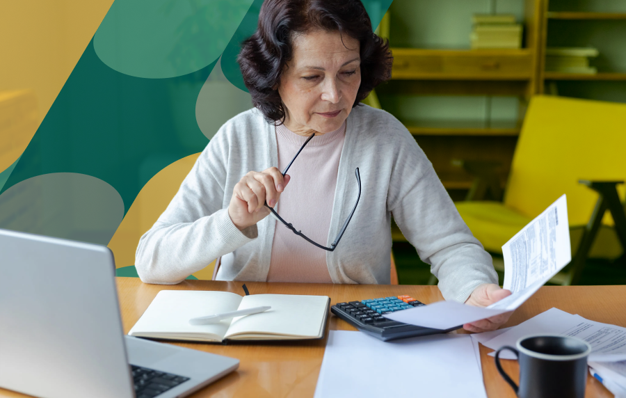 Close-up shot of a senior woman with wavy brown hair holding her glasses in one hand and sitting at a desk with many items in front of her - an open laptop, notebook and pen, calculator, stack of papers, and a mug.