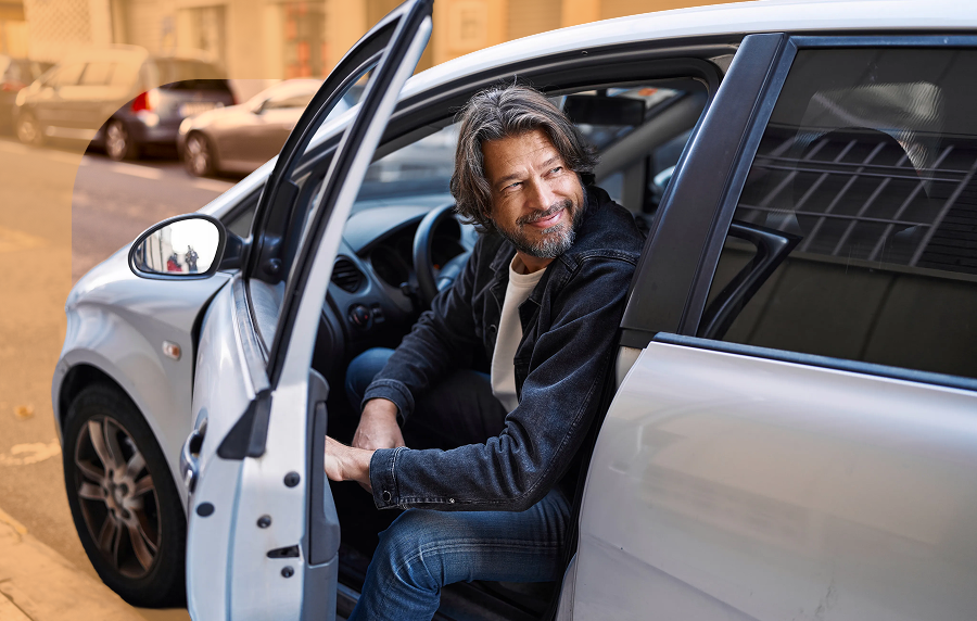 Middle aged man with facial hair wearing all denim getting out of the driver's seat of a silver car